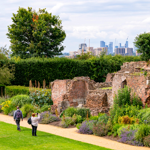Two adults stand beside the brick walls in the gardens at Eltham Palace with view of a section of London's skyline in the background.