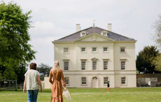 Two women walk towards the front of Marble Hill House in the gardens.
