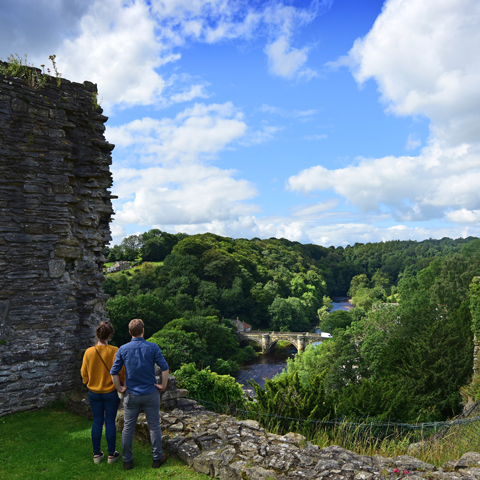 Two adults stand next to the stone wall ruins at Richmond Castle overlooking the view of a bridge and river.
