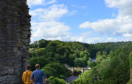 Two adults stand next to the stone wall ruins at Richmond Castle overlooking the view of a bridge and river.